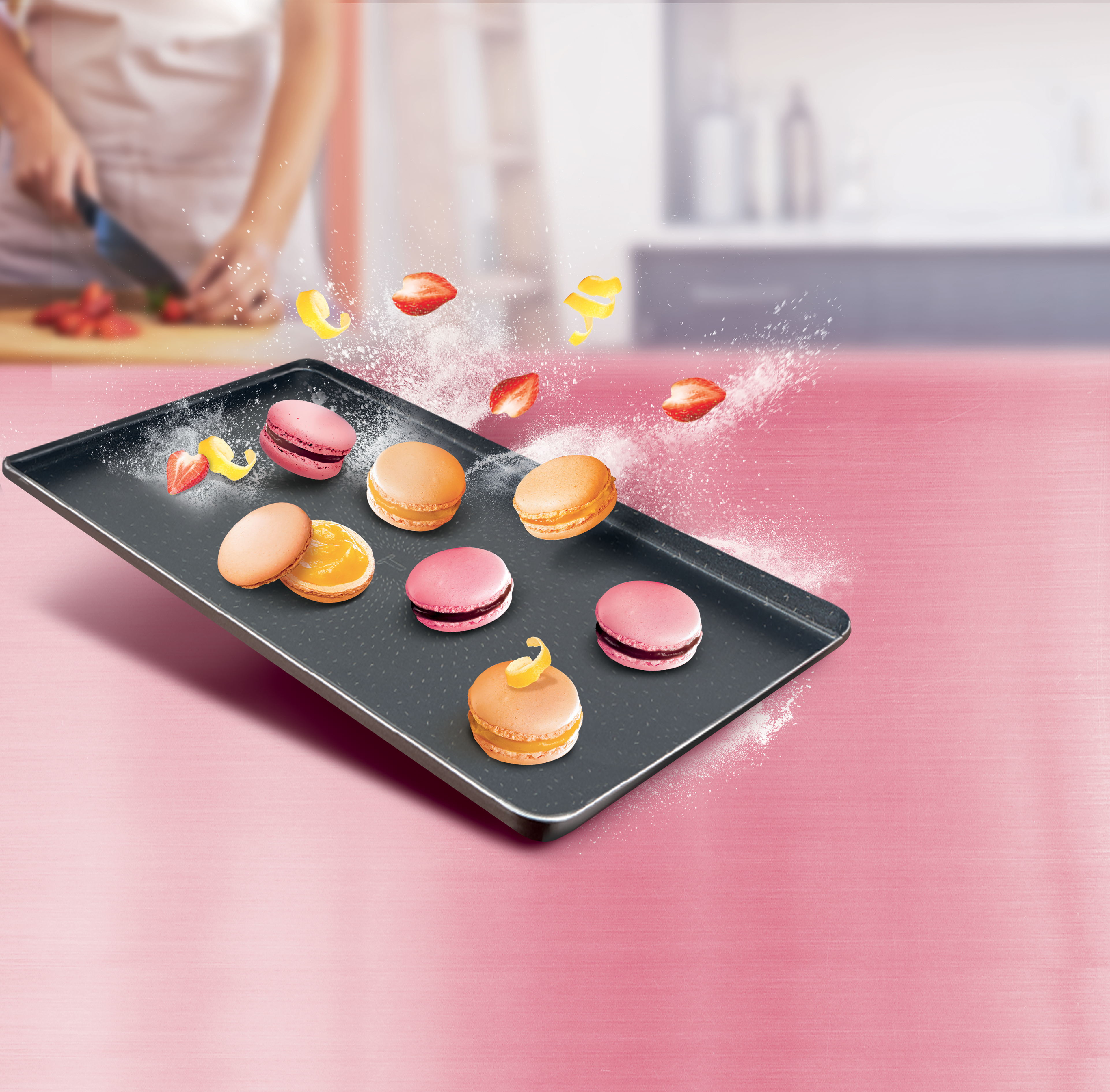 Egy tálca macaron, egy személy epret vág rózsaszín háttér előtt.