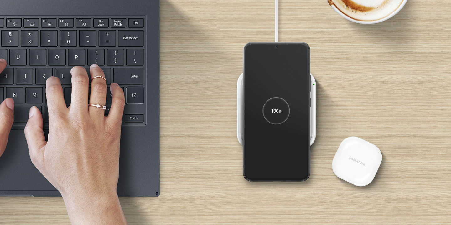 Woman's hands near laptop keyboard and phone charging on a pad, with a coffee.