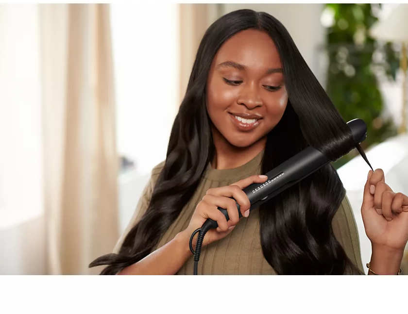 A woman with long, wavy hair using a black hair straightener, smiling.