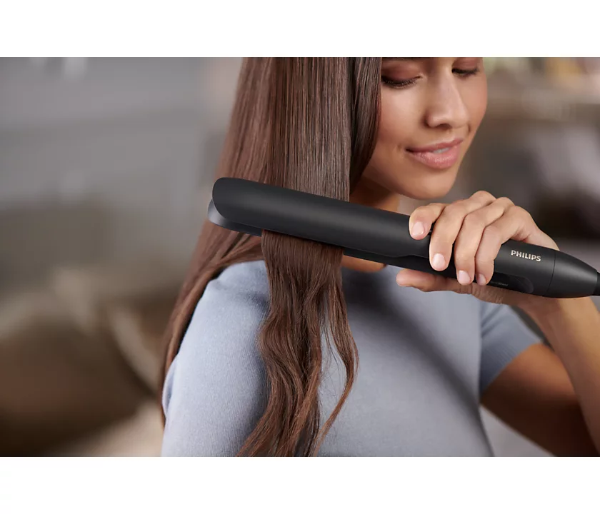 Woman using a black Philips hair straightener on her brown hair. She is smiling.