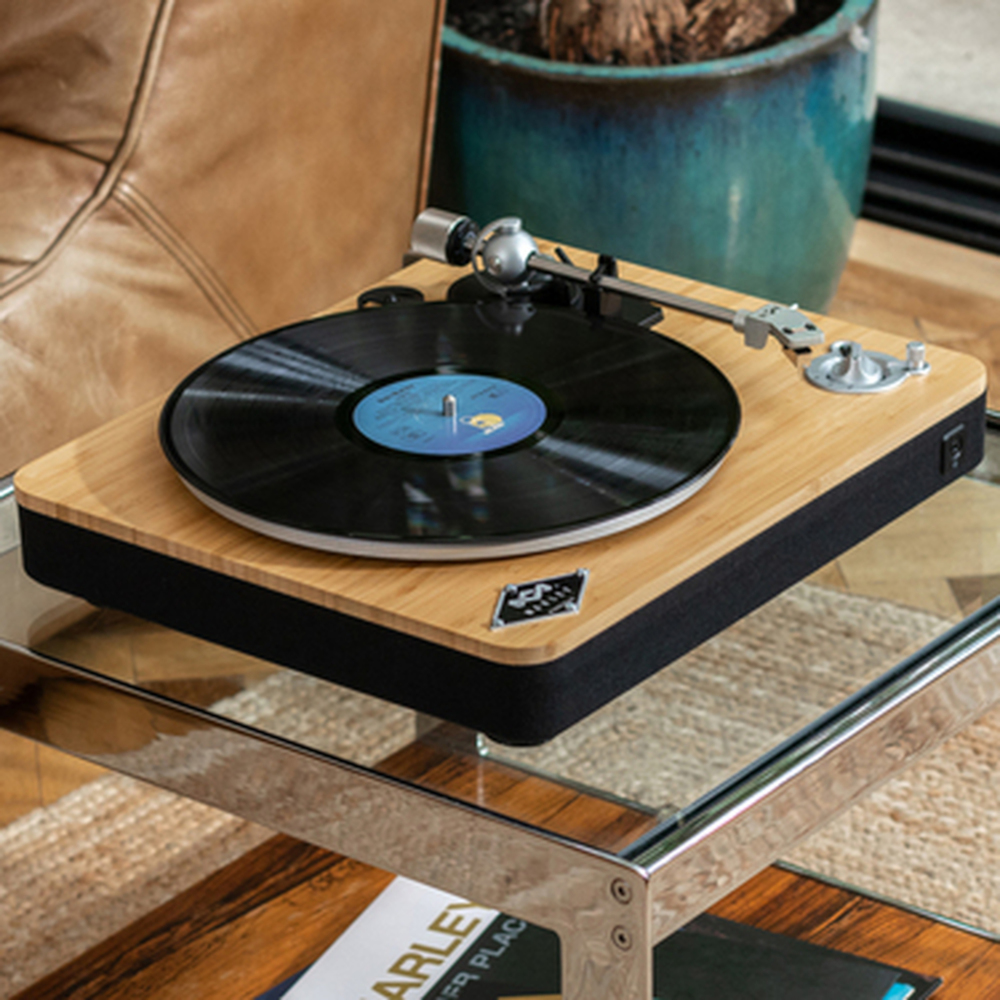 Een platenspeler op een glazen tafel. Een vinylplaat speelt, met een plant in een pot en een tijdschrift in de buurt.