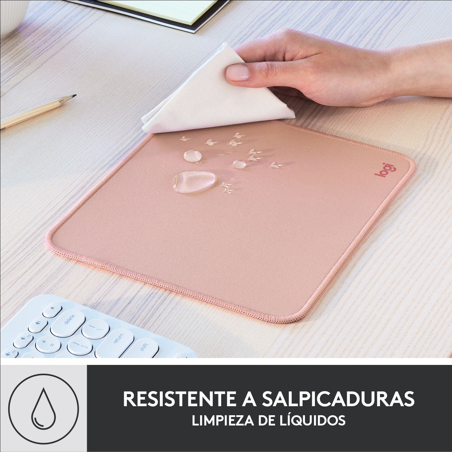 Una mano limpiando gotas de agua de una alfombrilla rosa con un paño blanco, un teclado y un logo.