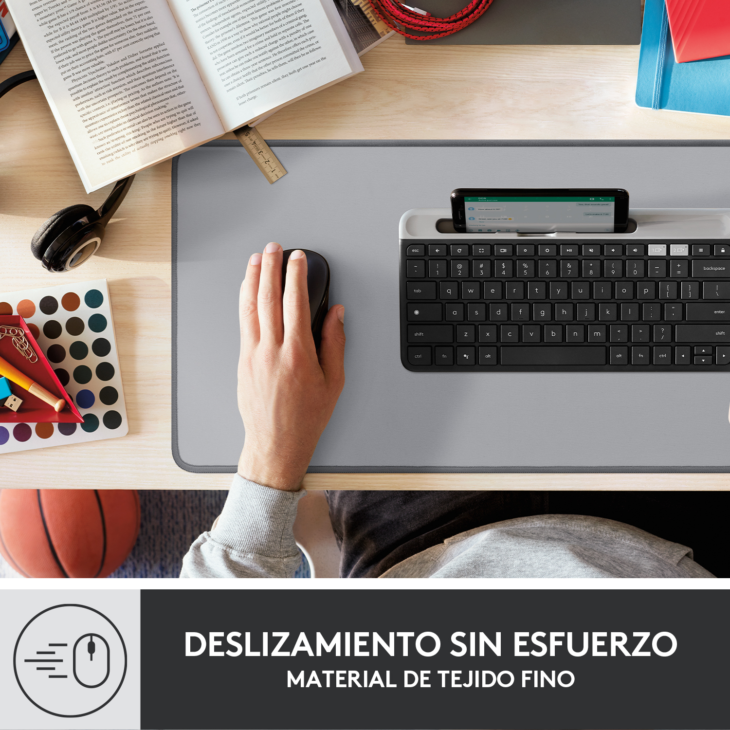 Una configuración de escritorio con un teclado, un ratón y un teléfono sobre una alfombrilla gris. Hay un libro y un bolígrafo en el escritorio.