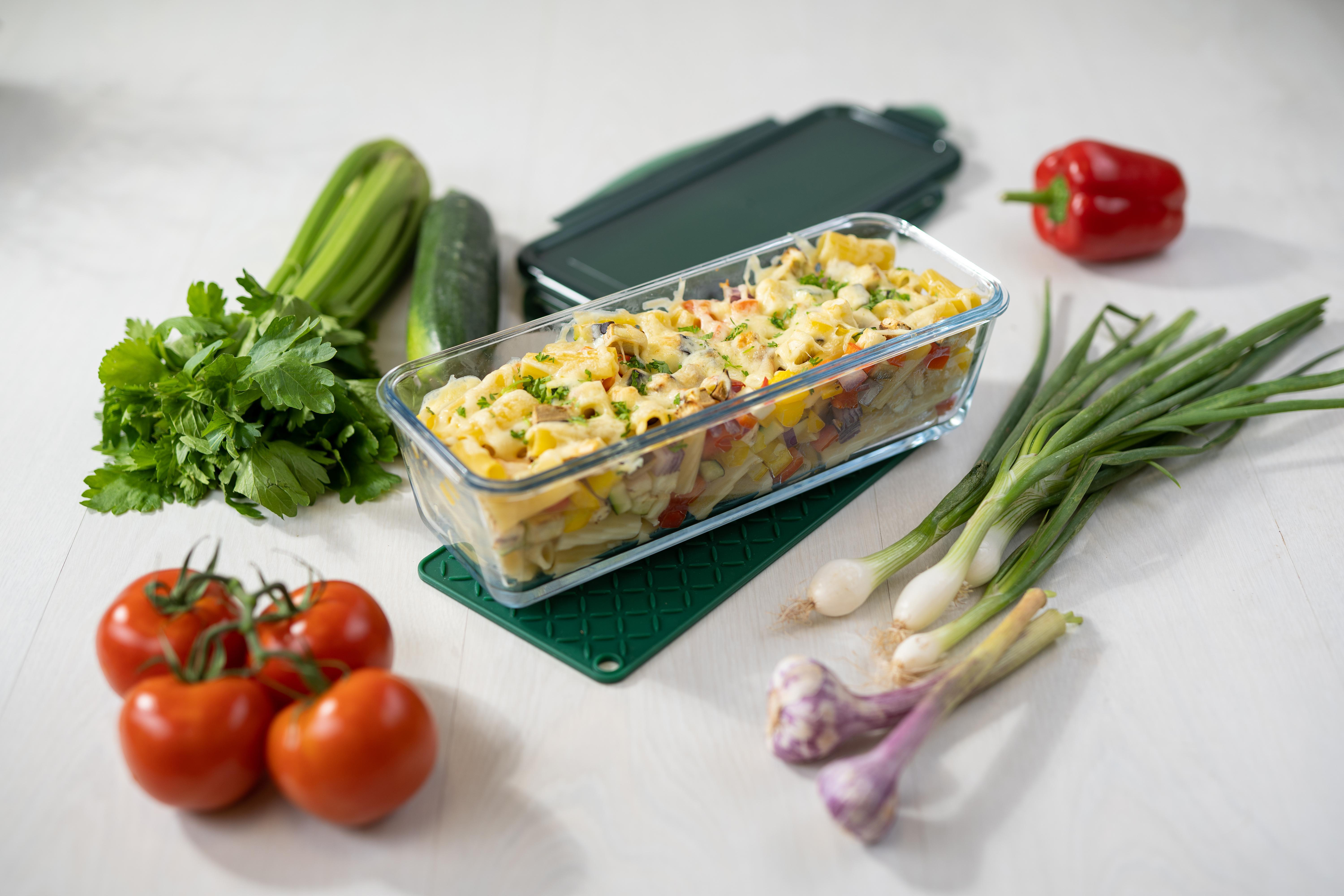 Eine Glasschüssel mit Pasta-Auflauf liegt auf einer grünen Matte mit Gemüse.