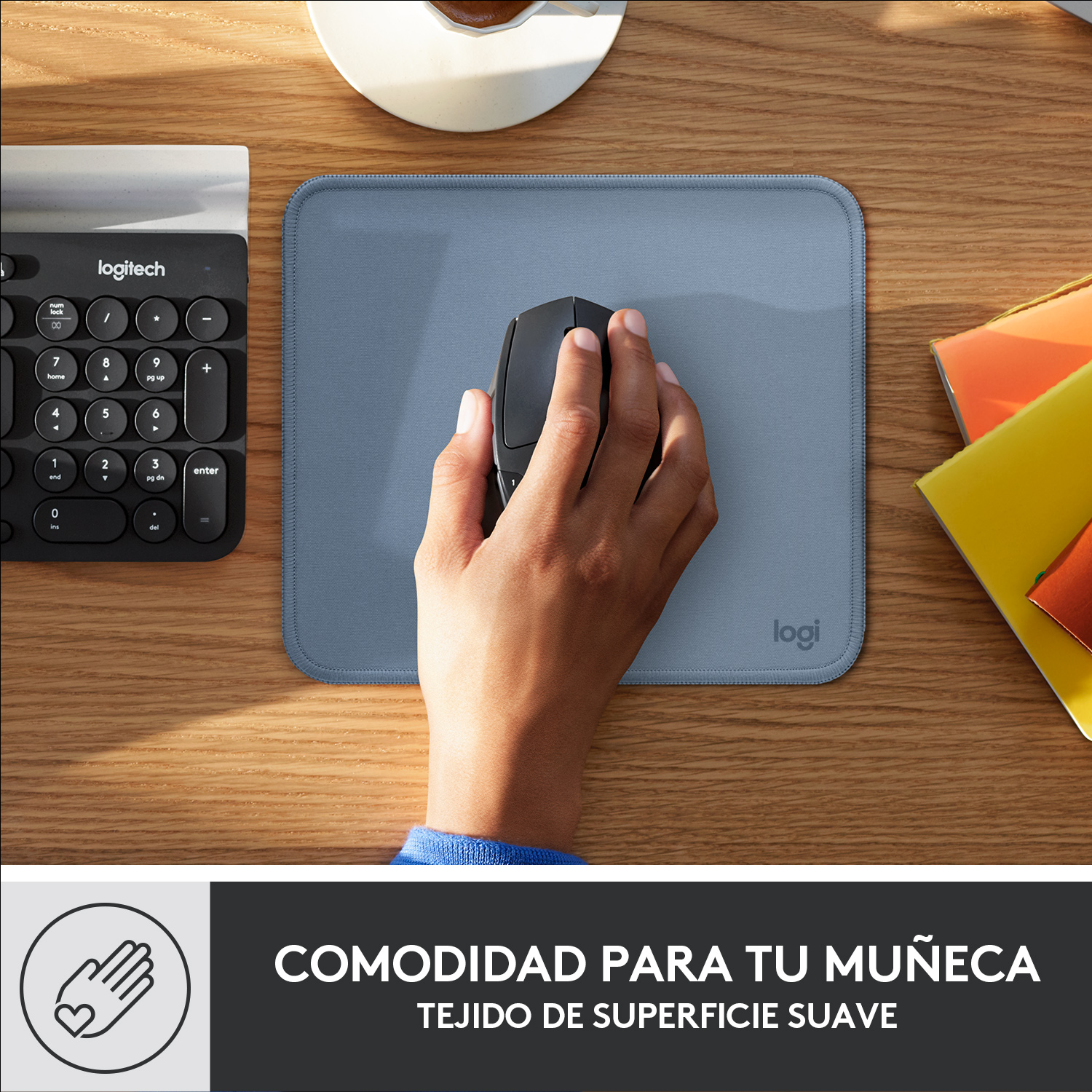 Mano usando un ratón en una alfombrilla azul. Teclado y cuadernos naranjas visibles.