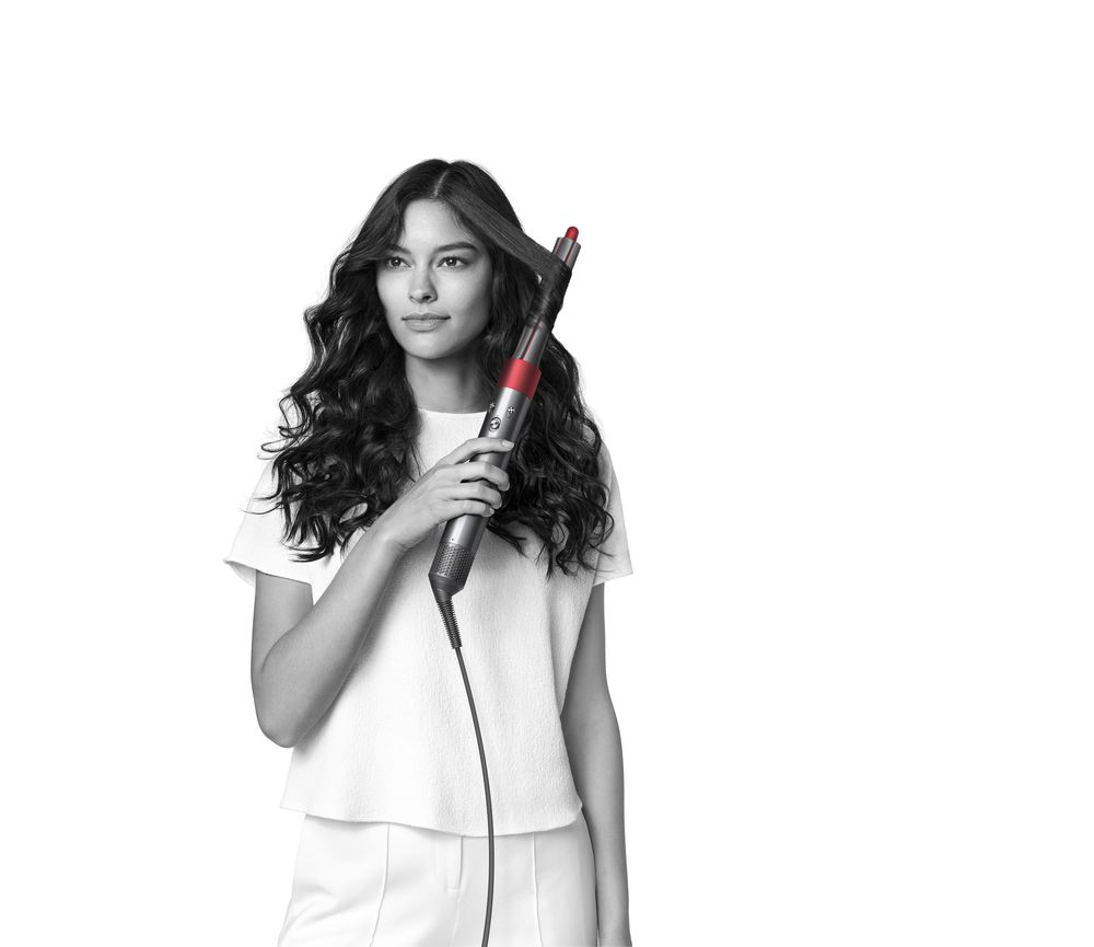 Een vrouw die een haarstylingtool gebruikt, met haar haar in golven gestyled, tegen een witte achtergrond.