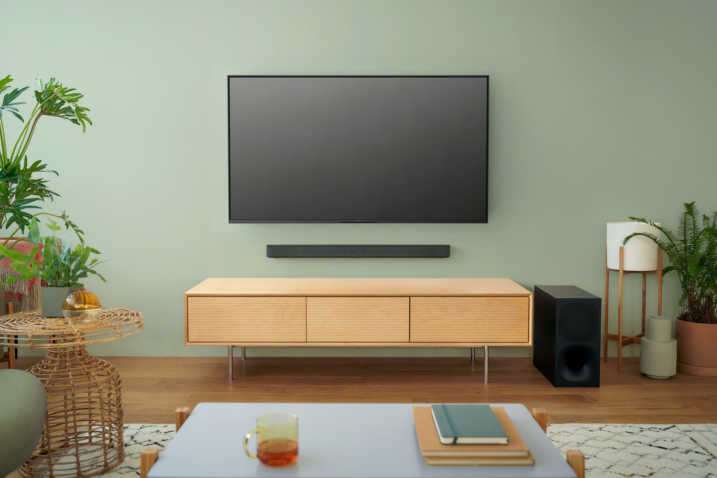Wohnzimmerszene: Fernseher an der Wand, Soundbar und Schrank. Pflanzen und ein Couchtisch im Vordergrund.