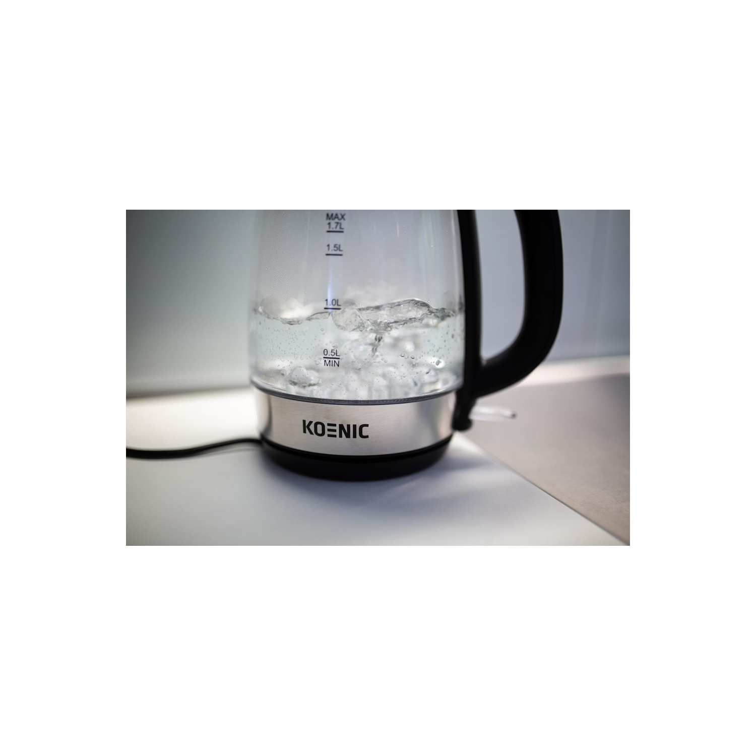 Hervidor eléctrico con agua hirviendo, base de acero inoxidable y asa negra. El vidrio transparente muestra niveles.