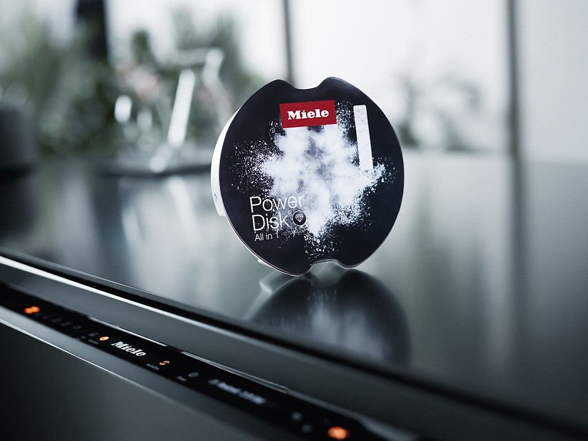 Ein Miele PowerDisk All in 1 Reinigungsmittel. Die Scheibe liegt auf einer Theke mit einem Miele-Gerät im Hintergrund.