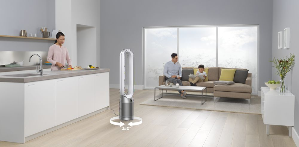 Una sala de estar con un ventilador, una mujer cocinando, una pareja en el sofá, ventana grande y paredes blancas.