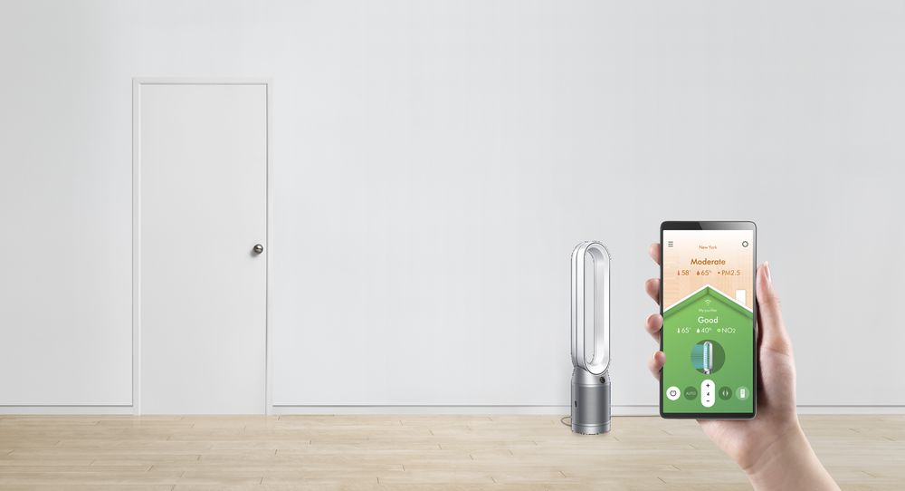 Un purificador de aire Dyson y una persona sosteniendo una tableta con lecturas de calidad del aire contra una pared blanca.
