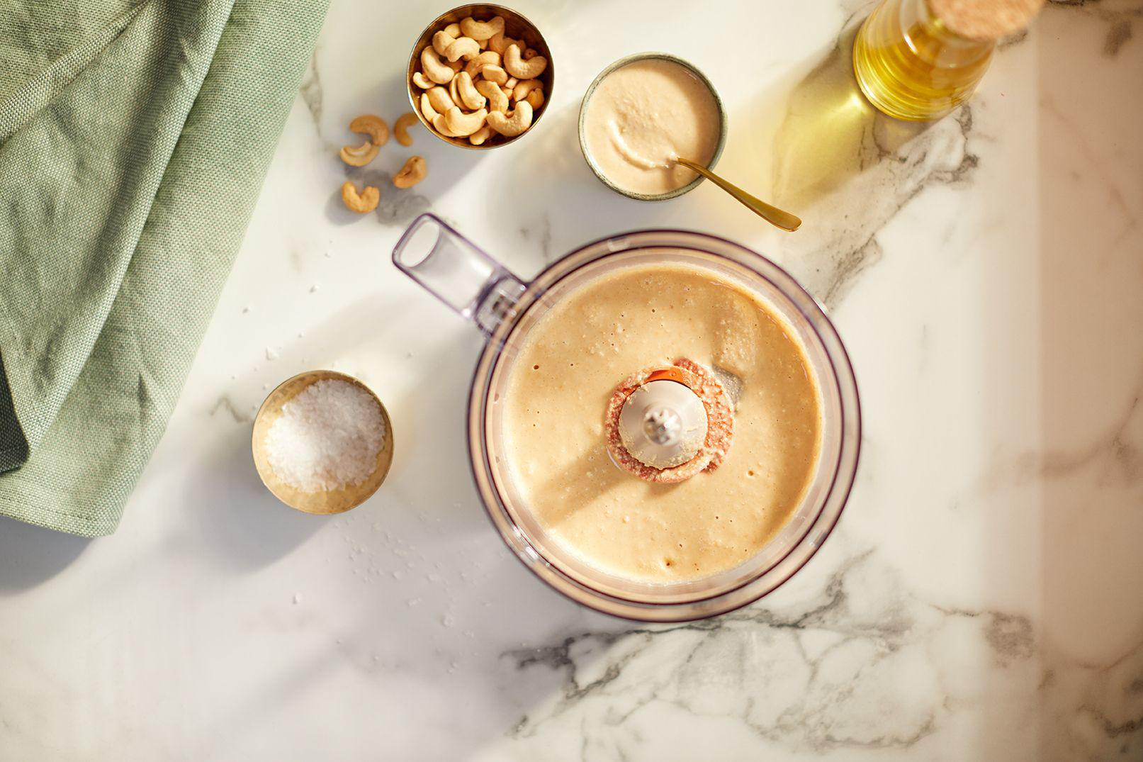 Küchenmaschine mit Cashewnüssen, Tahini und Olivenöl auf Marmoroberfläche.