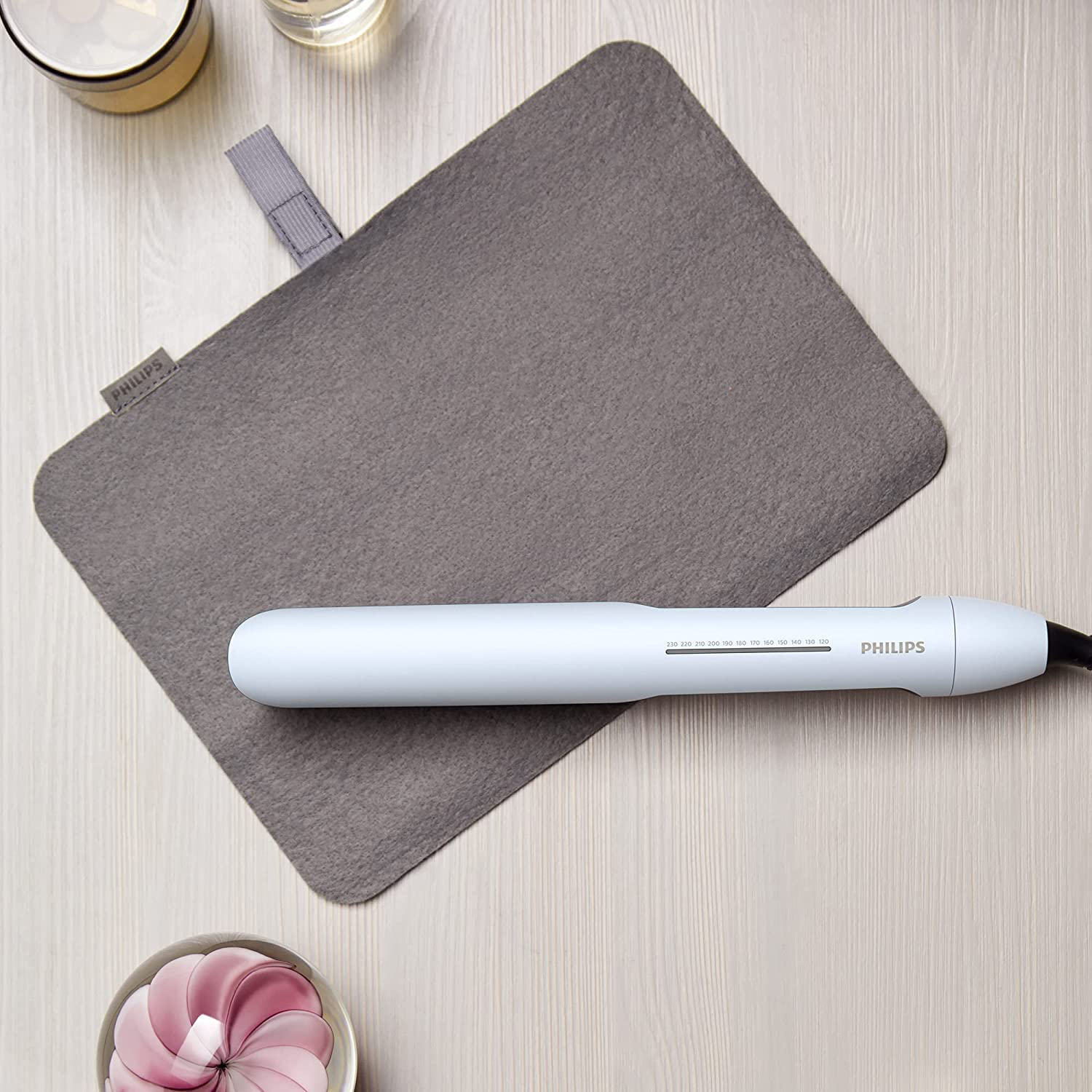 Una piastra per capelli Philips bianca su un tappetino in feltro grigio, accanto a piccoli oggetti su una superficie di legno chiaro.