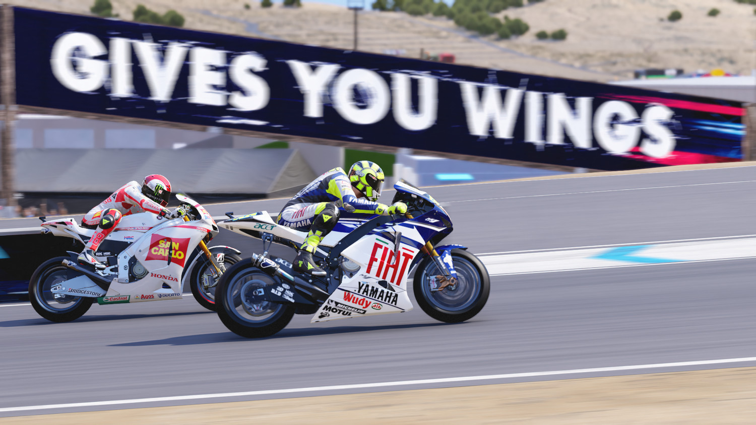 Deux motos en course sur une piste. Une bannière dit "Gives You Wings".