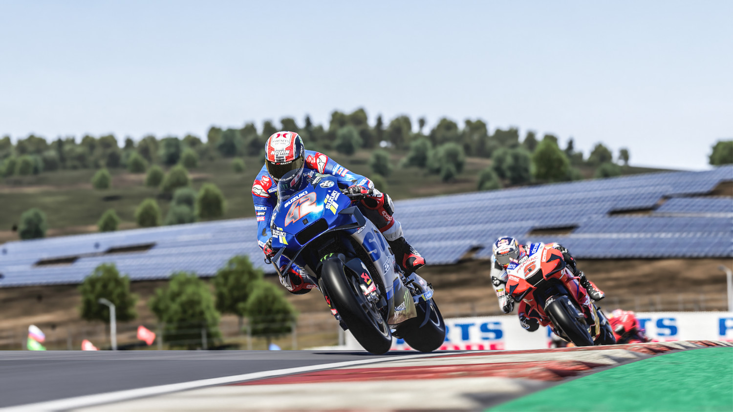 Pilotes de moto sur une piste. Un pilote en Suzuki bleu, l'autre en rouge. Arbres verts et ciel en arrière-plan.