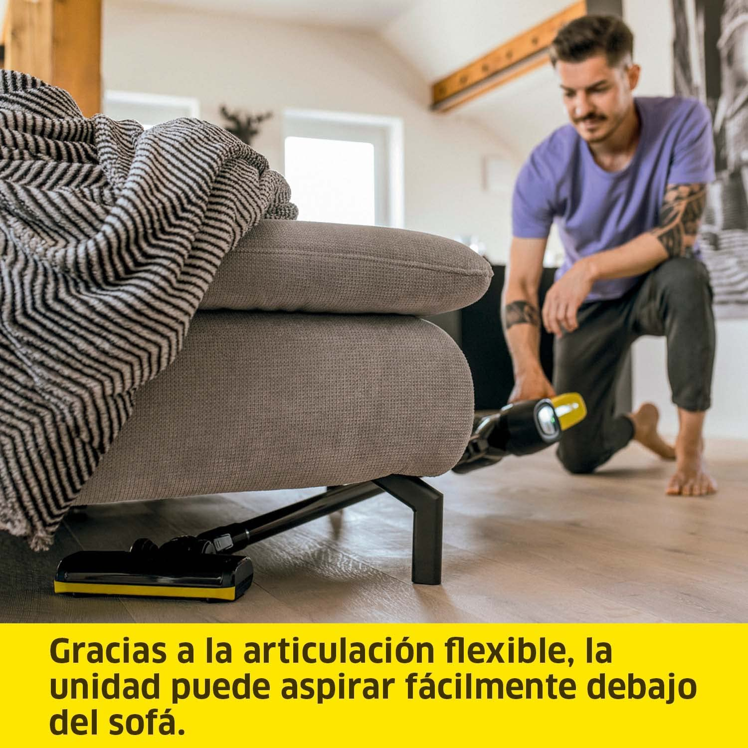 Hombre aspirando debajo de un sofá. La aspiradora tiene cabezal amarillo y negro.