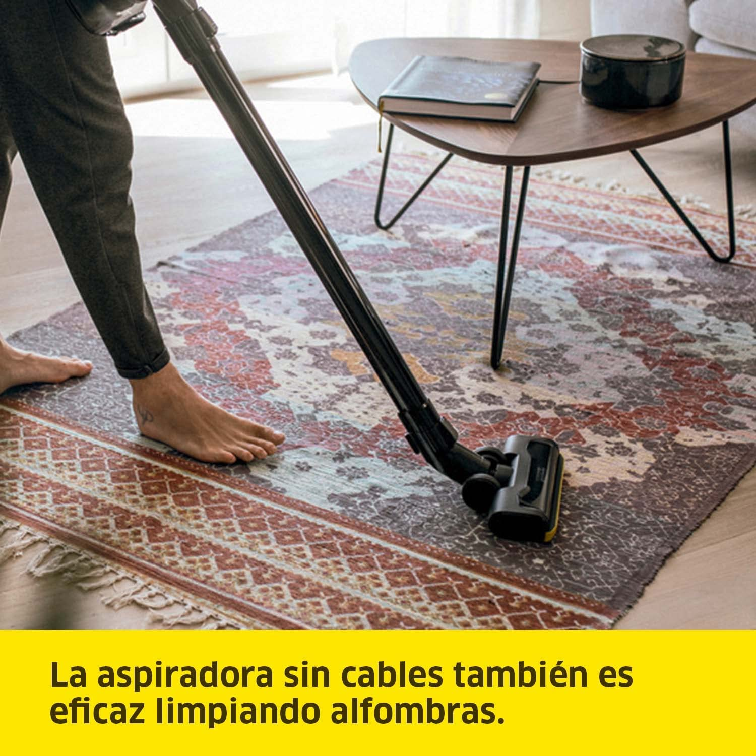 Una persona aspirando una alfombra estampada con una aspiradora inalámbrica. Una mesa está en el fondo.
