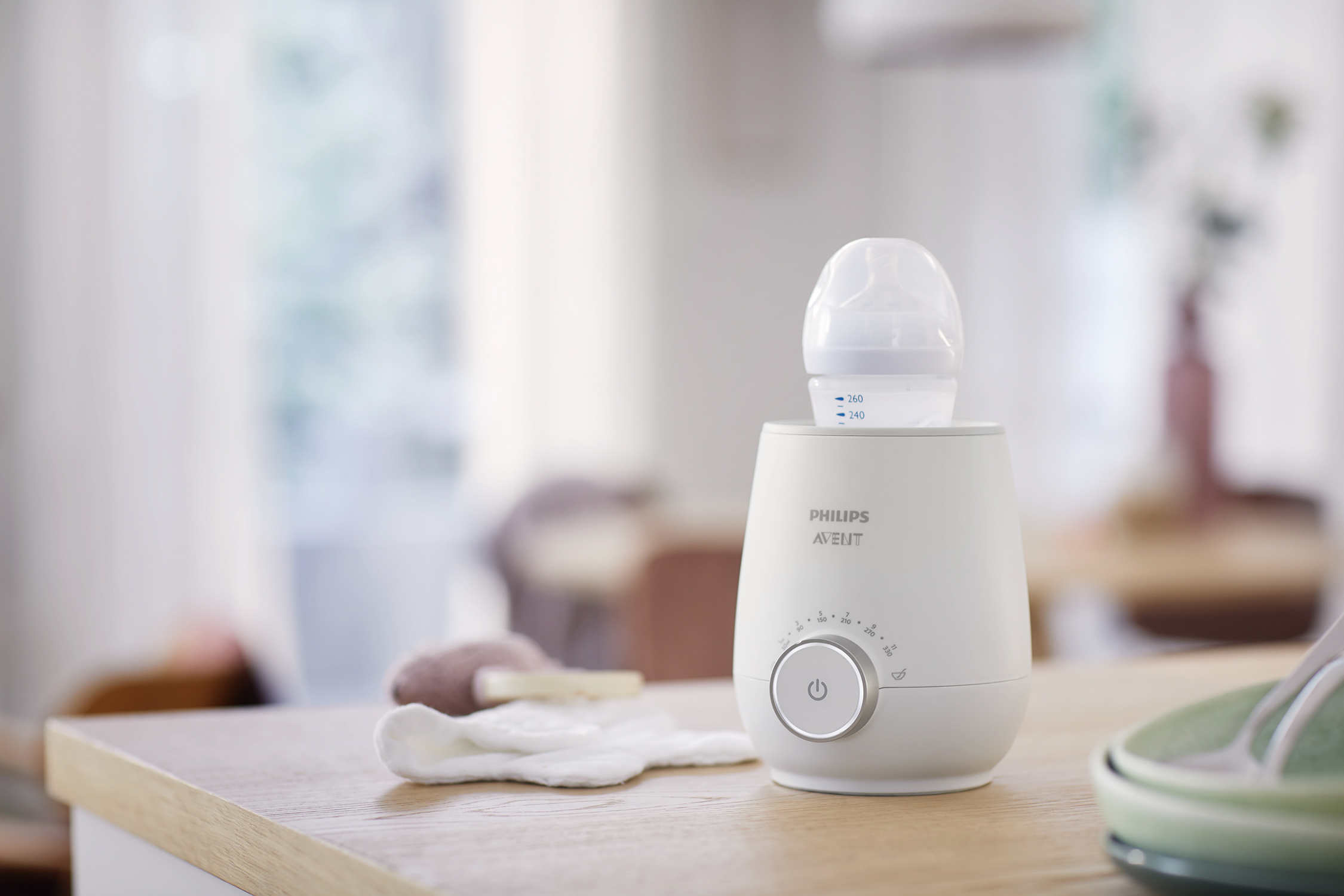 Een witte babyflessenwarmer met een fles erin. Een doek en een flessenborstel liggen aan de zijkant.