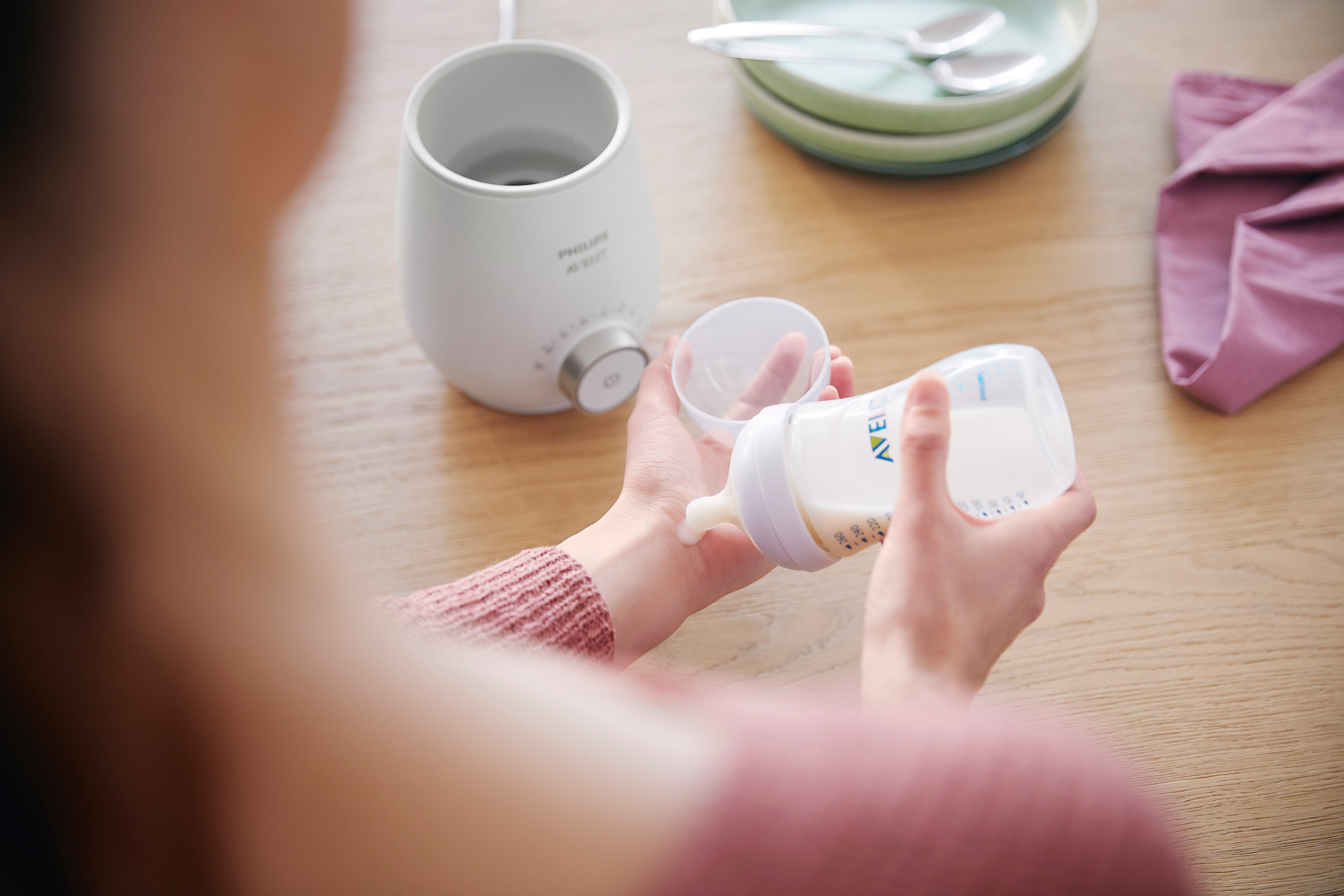 Een persoon giet melk van een fles in een andere fles. Er staat een flessenwarmer links.