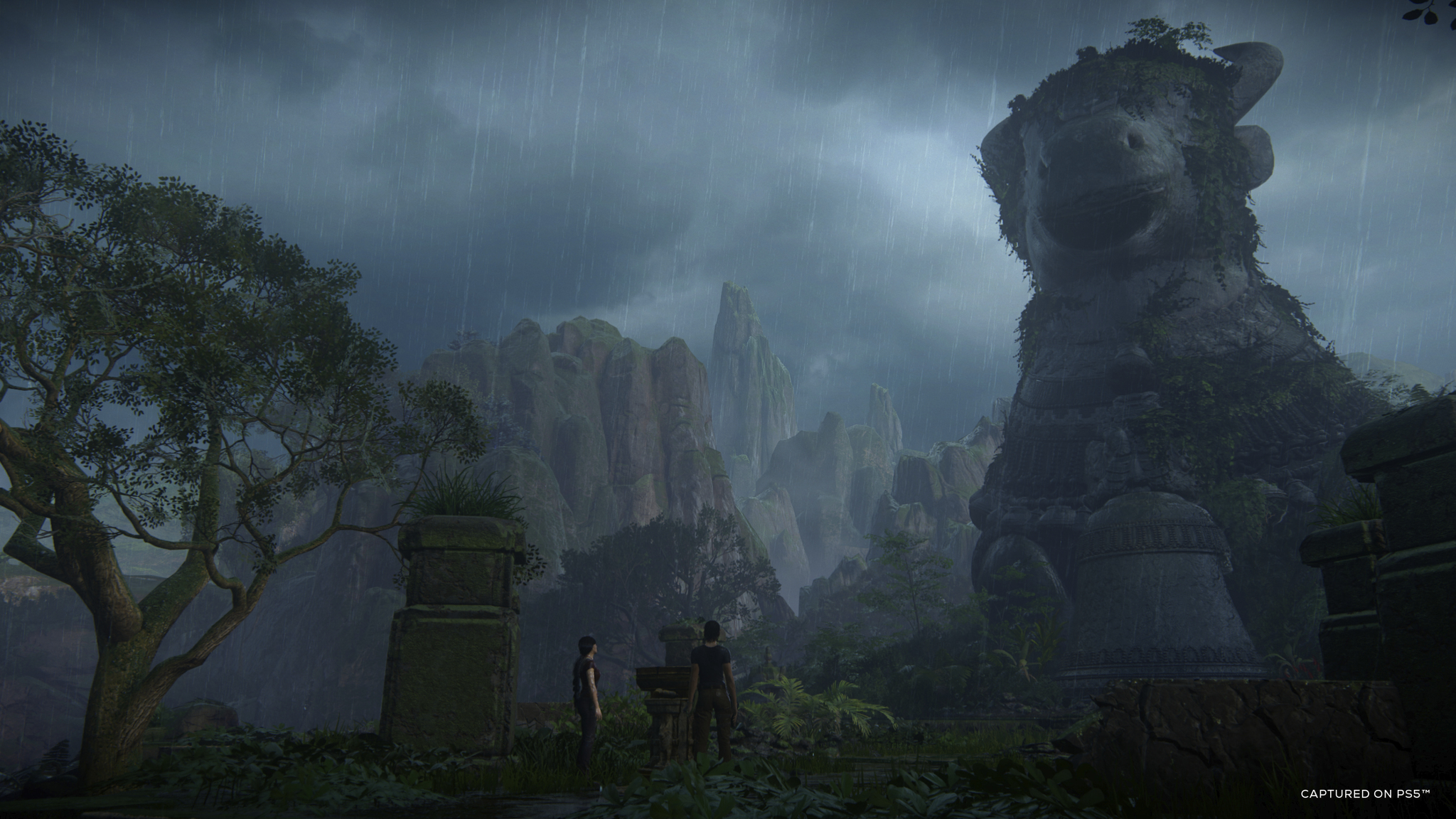 Two figures in a forested area with a giant statue and background mountains. Overcast sky.