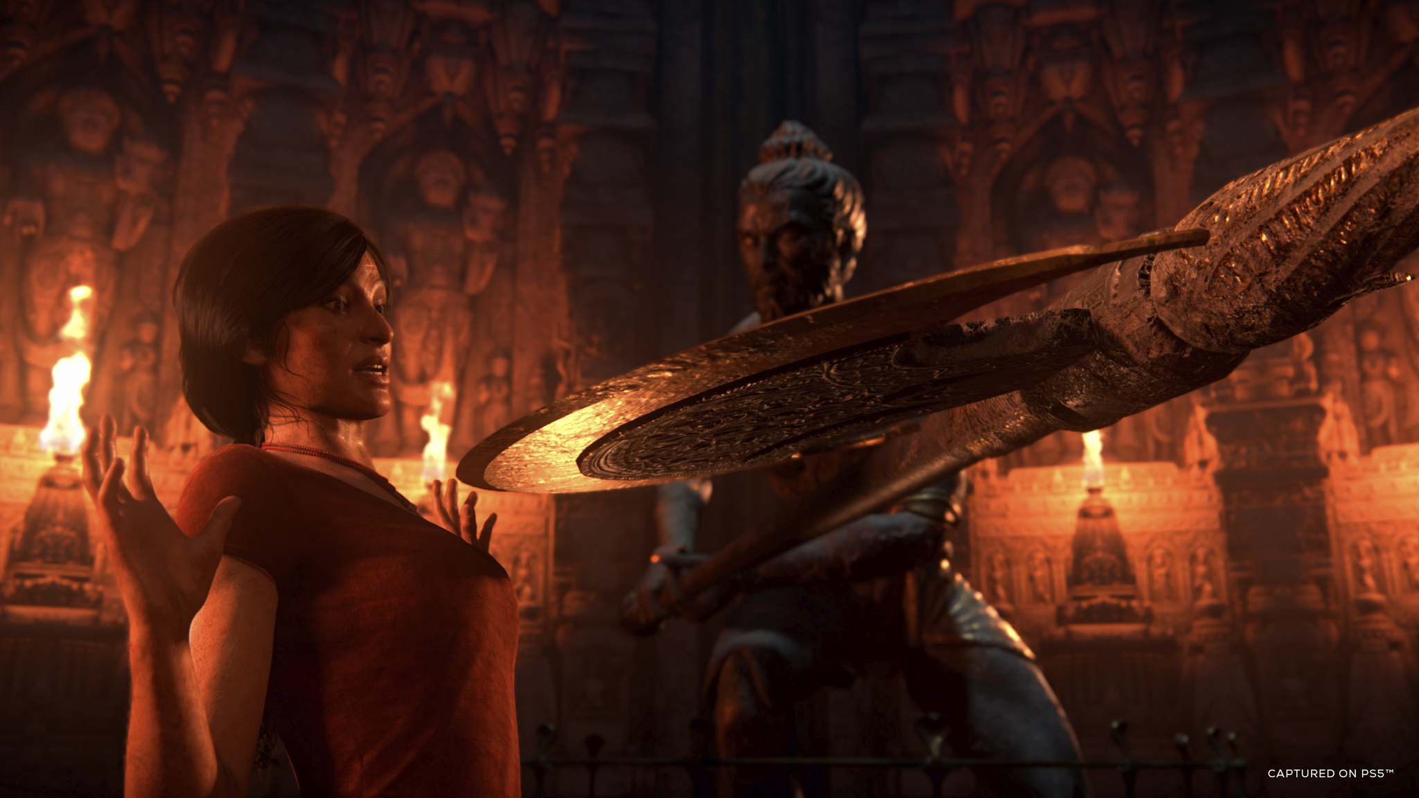 Woman facing a statue with a large weapon in a temple. The scene is lit with fire.