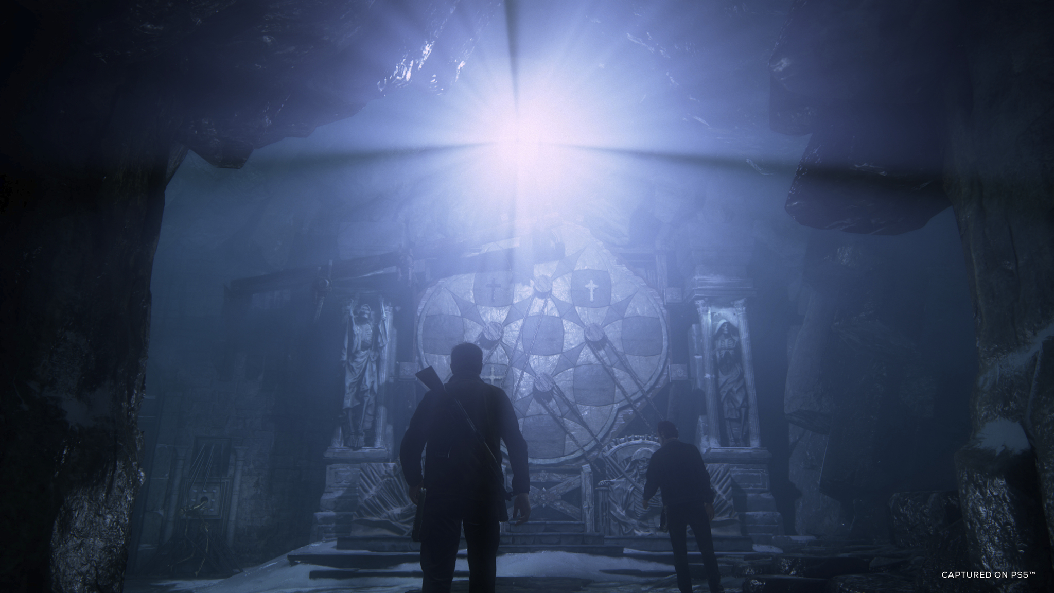 Two figures stand before a large, ornate stone structure in a cavern. Bright light shines from above. Statues flank the structure.