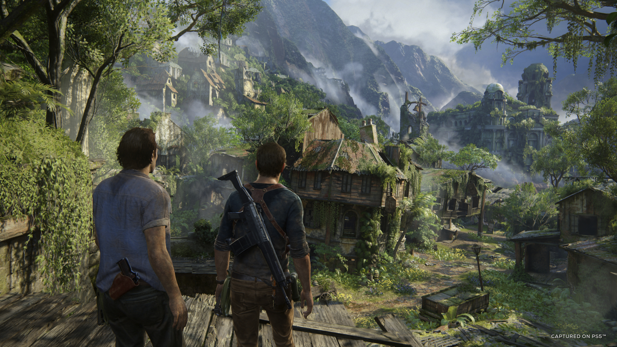 Two men looking at a village with green trees and mountains in the background.