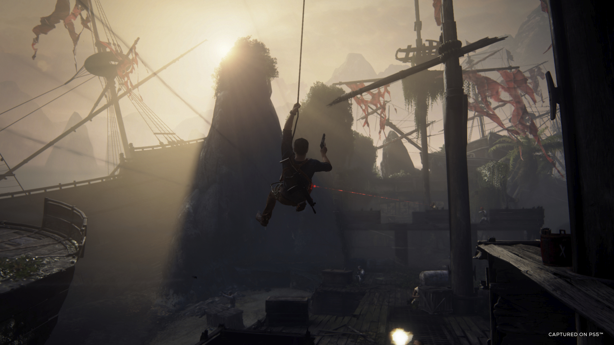 Man hanging from a rope, swinging over water, with a boat and mountains in the background. Sunlit scene.