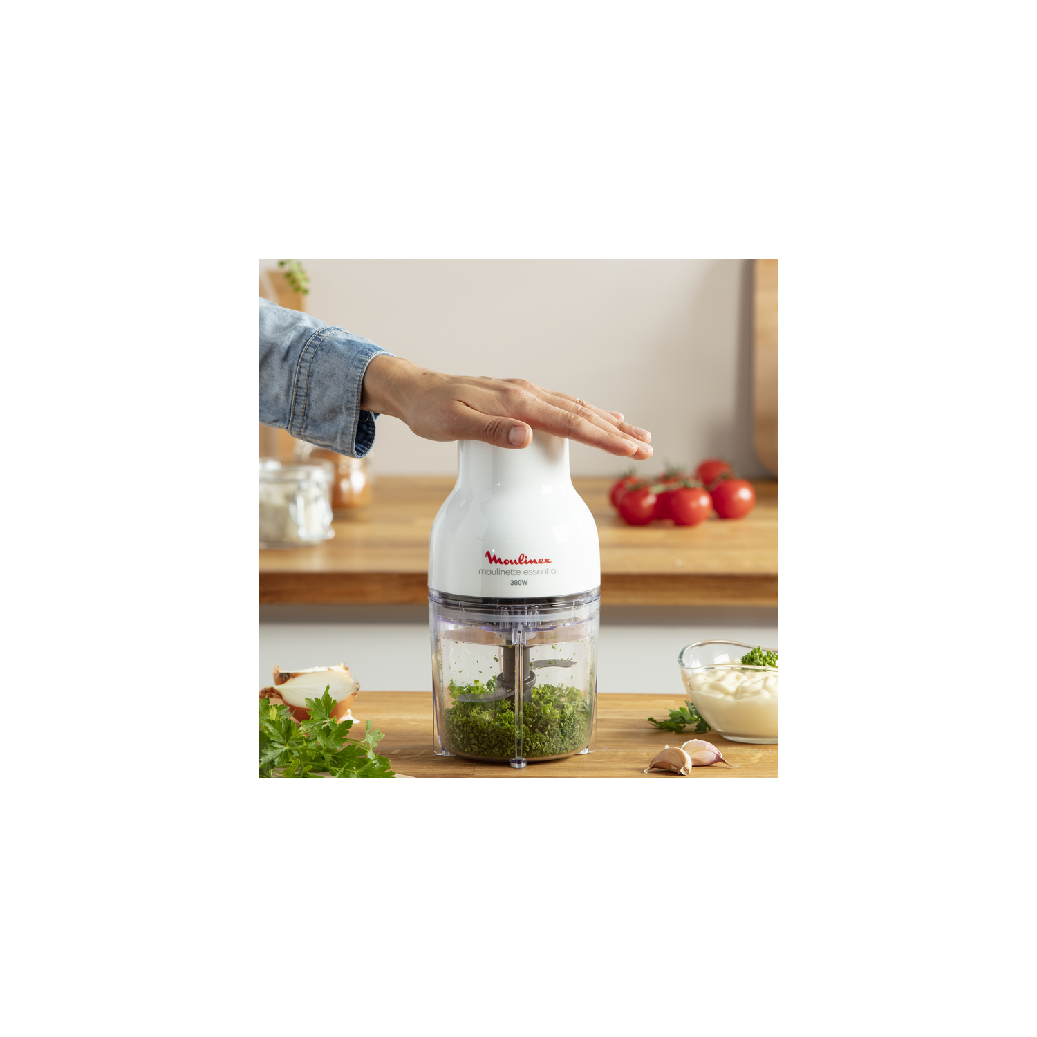 Una mano presiona un picador de alimentos Moulinex blanco, que contiene hierbas y verduras picadas.
