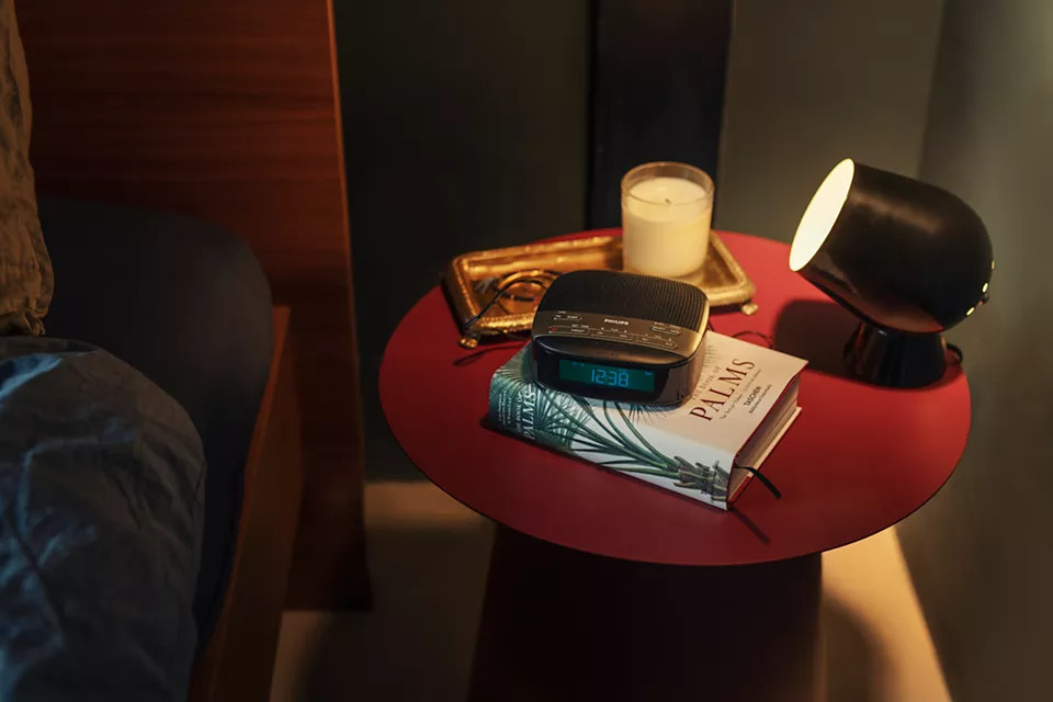 Scène de chambre avec une table de chevet rouge, un radio-réveil et une lampe allumée.