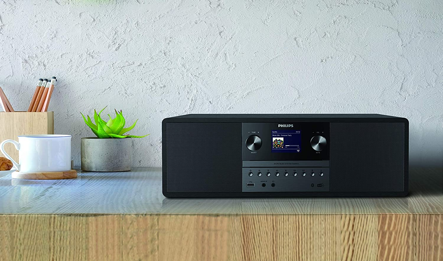 Een Philips stereosysteem staat op een houten oppervlak. Er zijn ook een witte mok, potloden en een plant in een betonnen pot aanwezig.