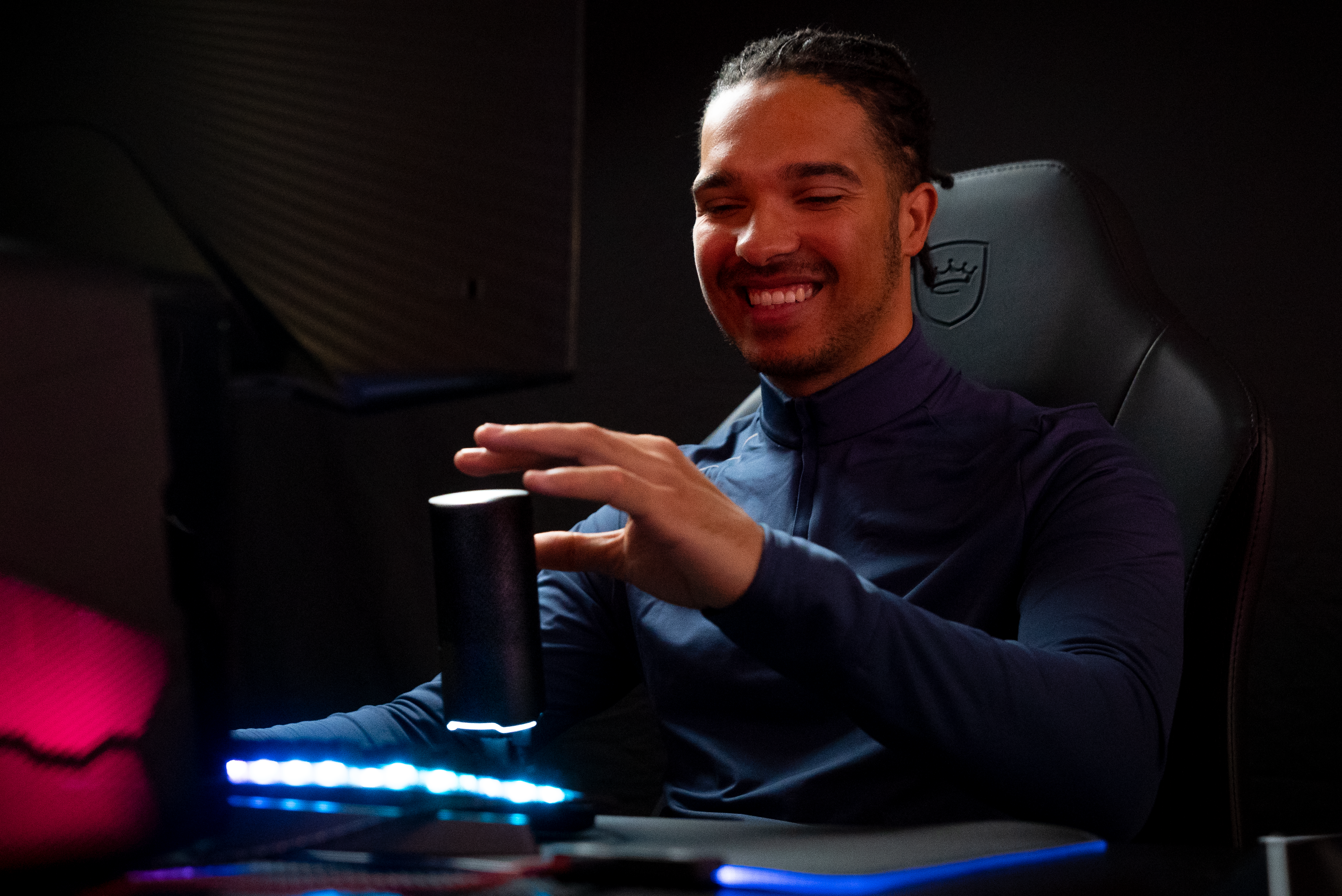 Mann lächelt, hält ein schwarzes zylindrisches Objekt, Gaming-Setup im Hintergrund.