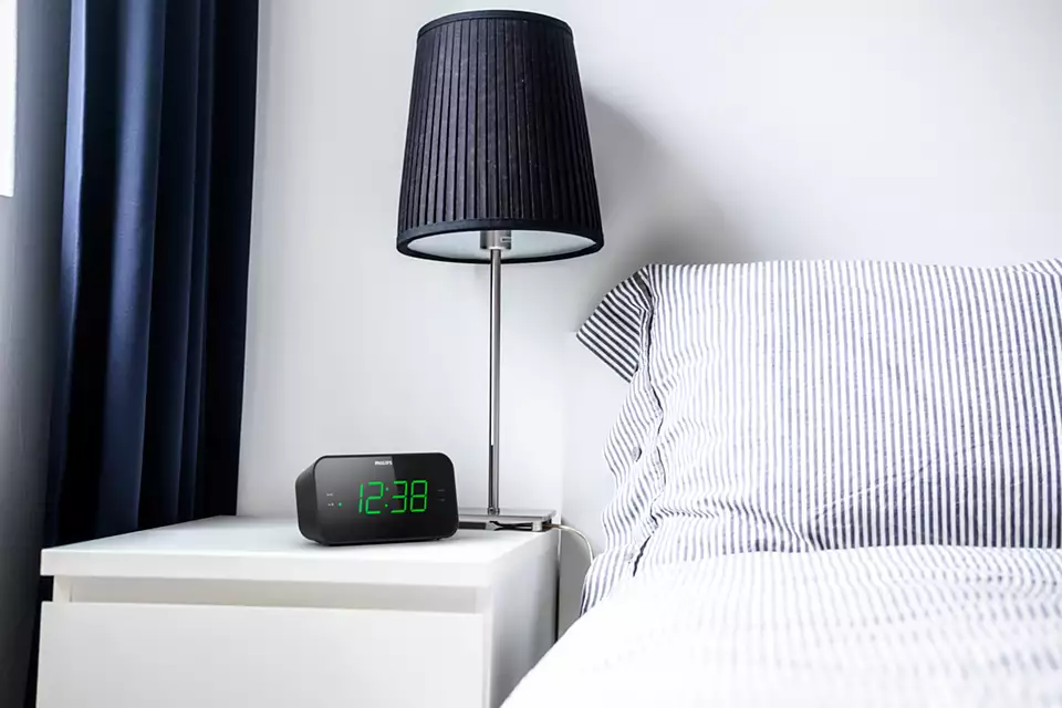 Un réveil Philips noir sur une table de chevet blanche à côté d'un lit avec une literie à rayures.