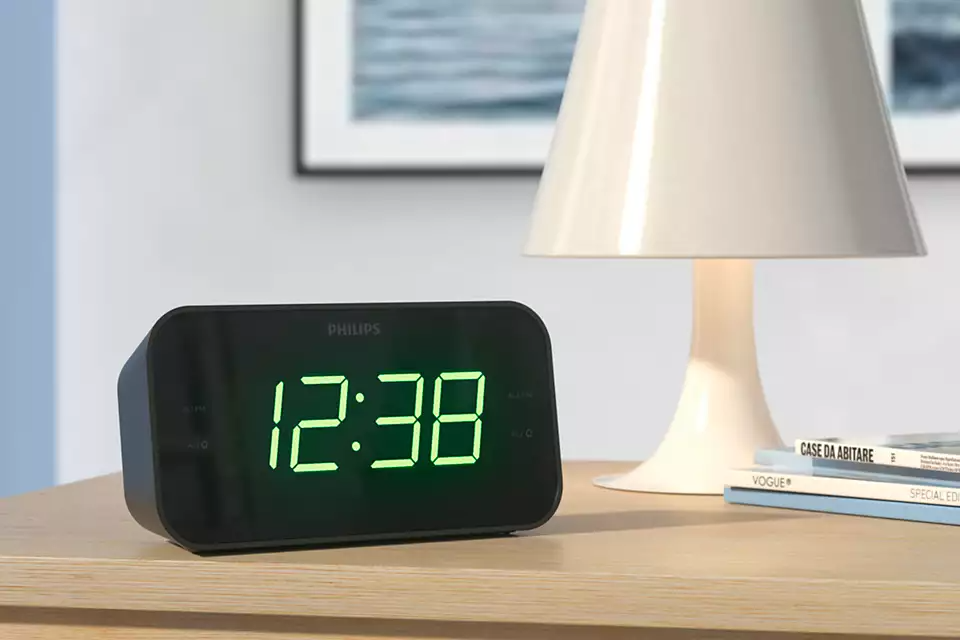 L'horloge numérique noire affiche 12:38 en chiffres verts. Une lampe blanche à droite et une peinture derrière.