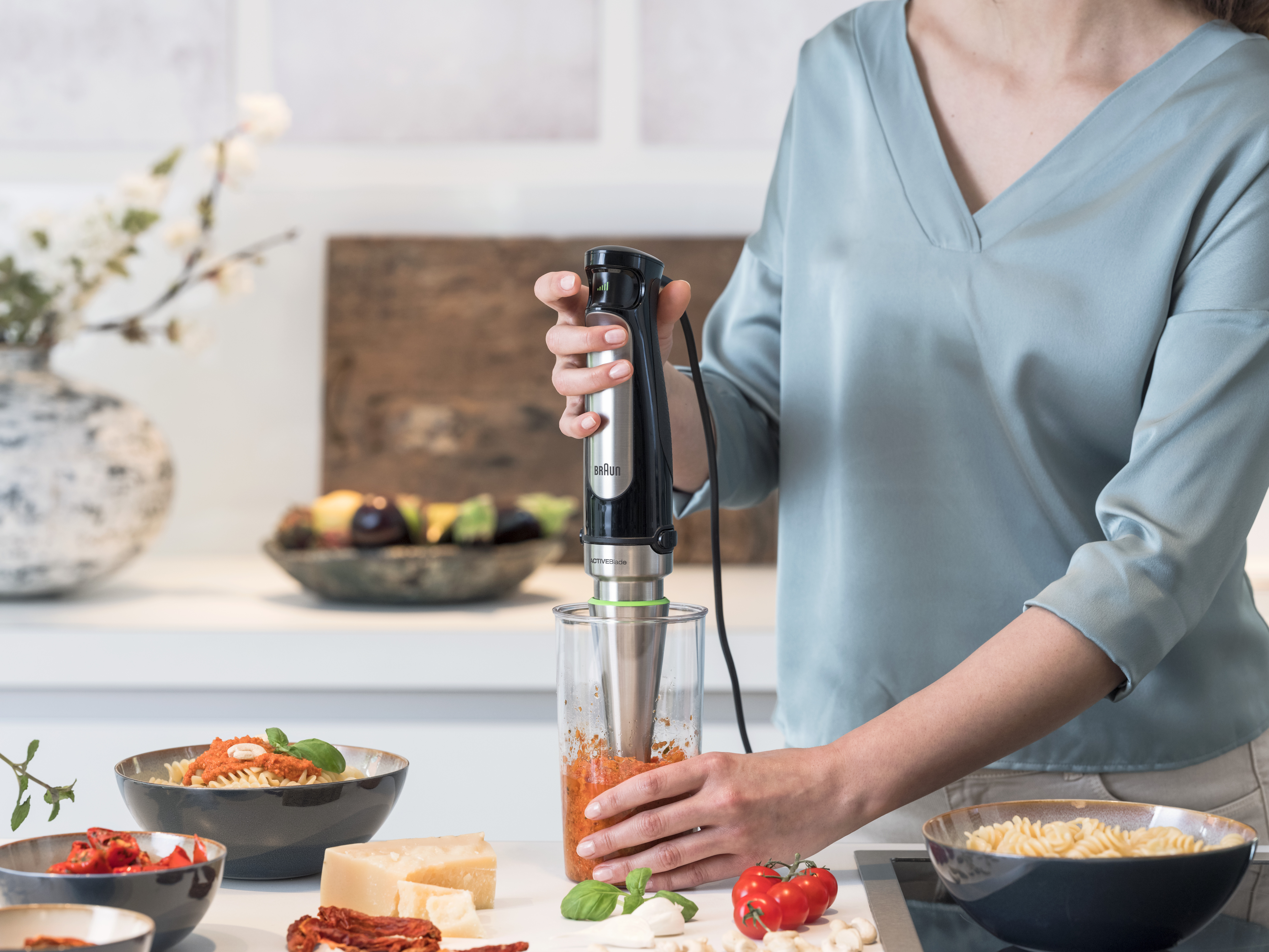 Woman using a silver and black hand blender over a glass container.
