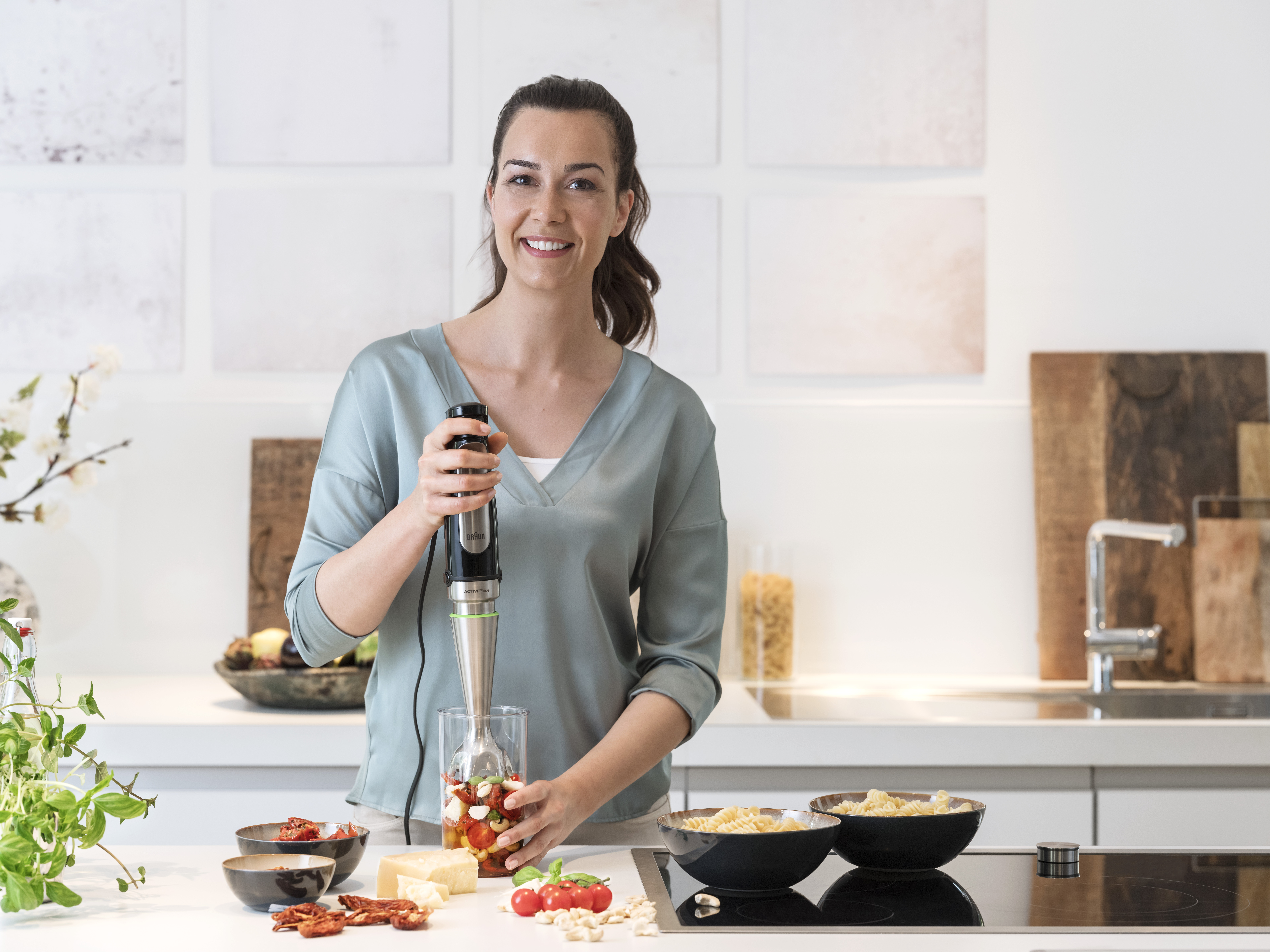 A woman smiling, using an immersion blender in a glass with tomatoes and other ingredients.