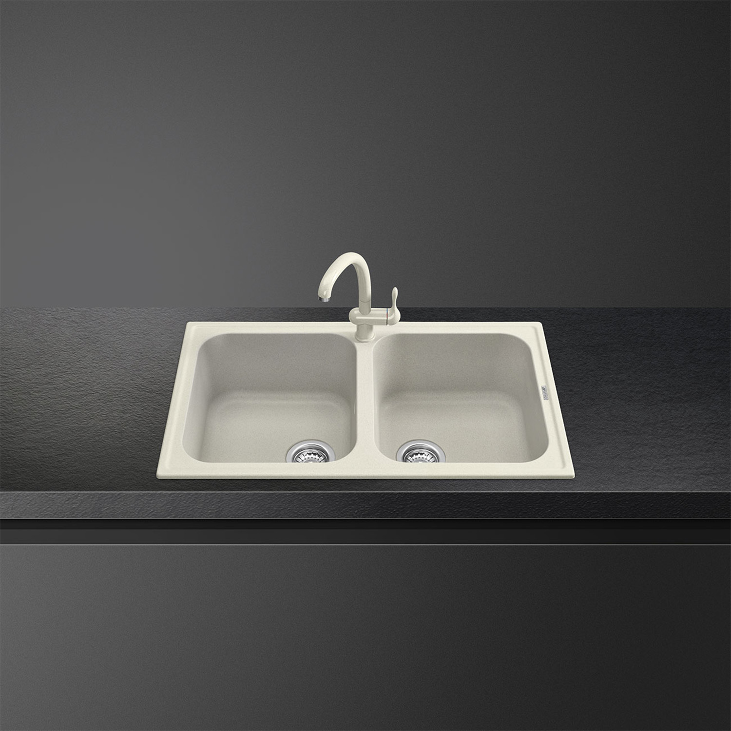 Lavello doppio color crema con rubinetto su ripiano scuro. Lo sfondo è un muro scuro.