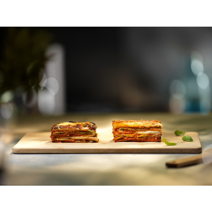 Két négyzet lasagne egy fa deszkán, bazsalikom levelekkel mellette.