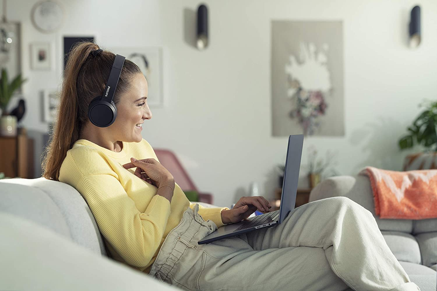 Vrouw met koptelefoon gebruikt laptop op een bank. Helder, gezellig interieur.
