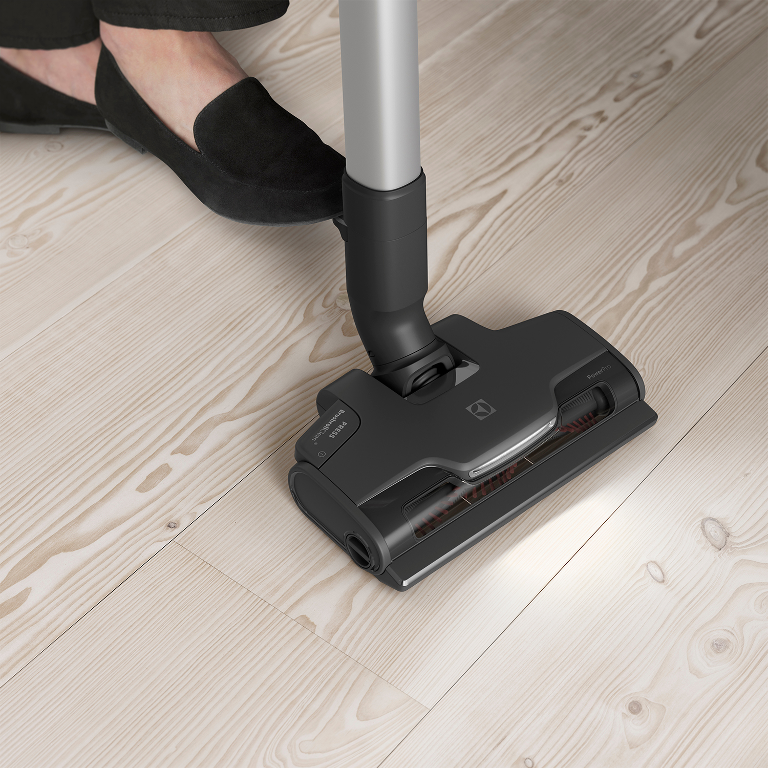 Testa dell'aspirapolvere su pavimento in legno chiaro con piede di una persona e scarpa scura sullo sfondo.