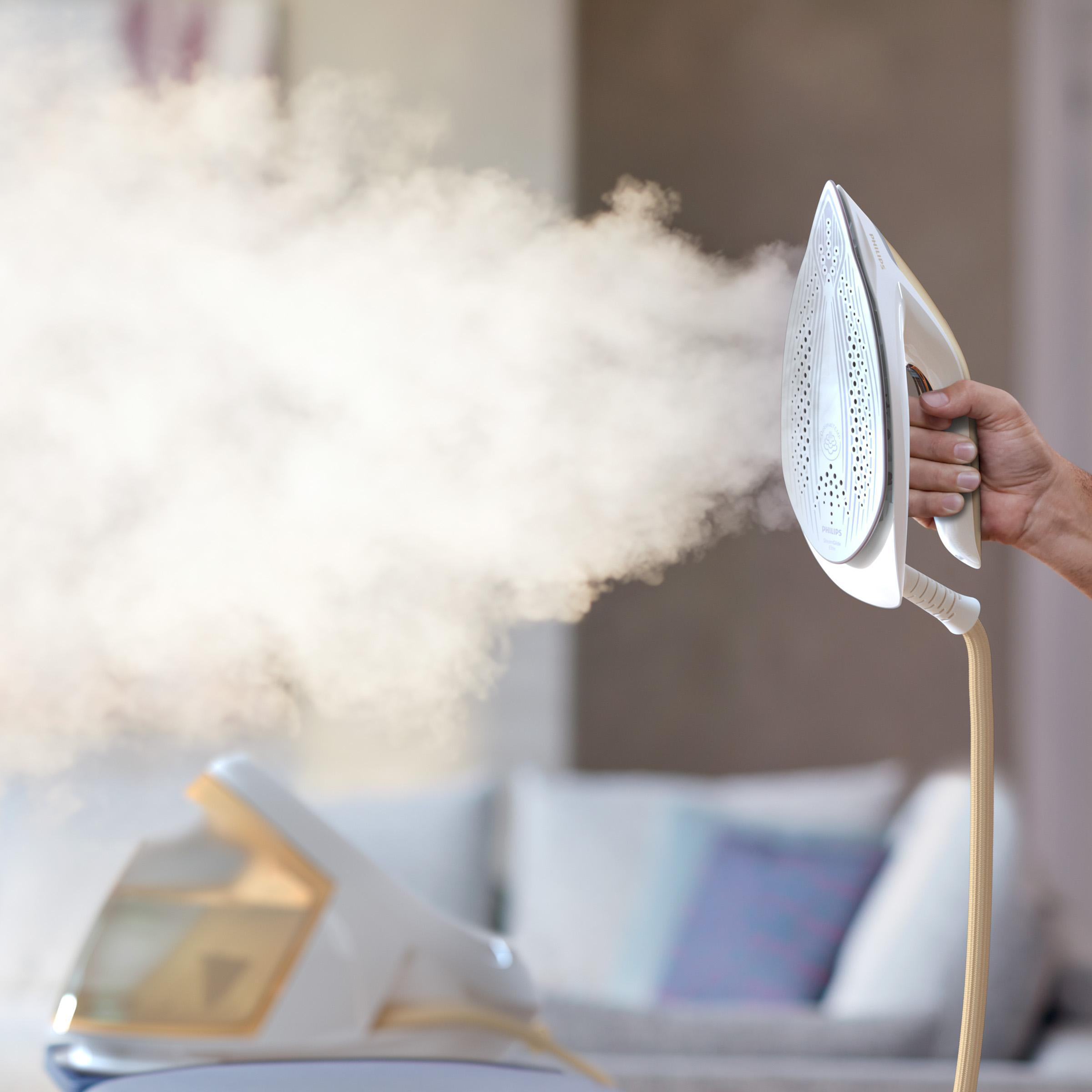 Ein weißes und goldenes Bügeleisen dampft. Eine Hand hält das Bügeleisen, Dampf kommt heraus, und eine Basis ist im Hintergrund.
