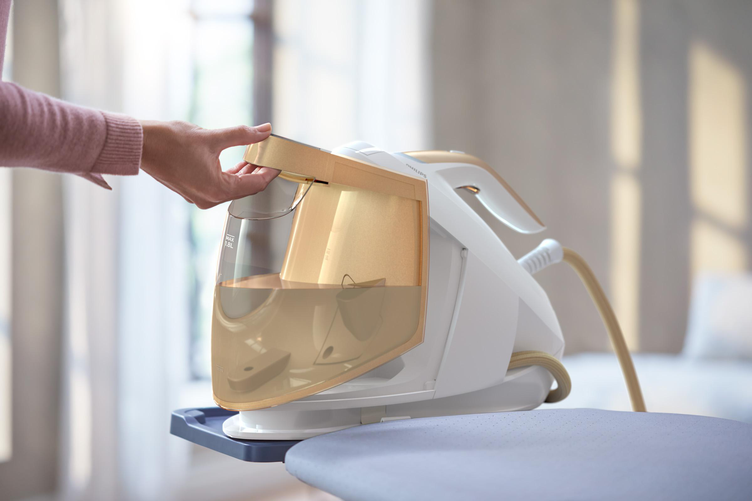 Eine Hand öffnet einen Wassertank auf einem weiß-goldenen Dampfbügeleisen, das auf einem Bügelbrett steht.