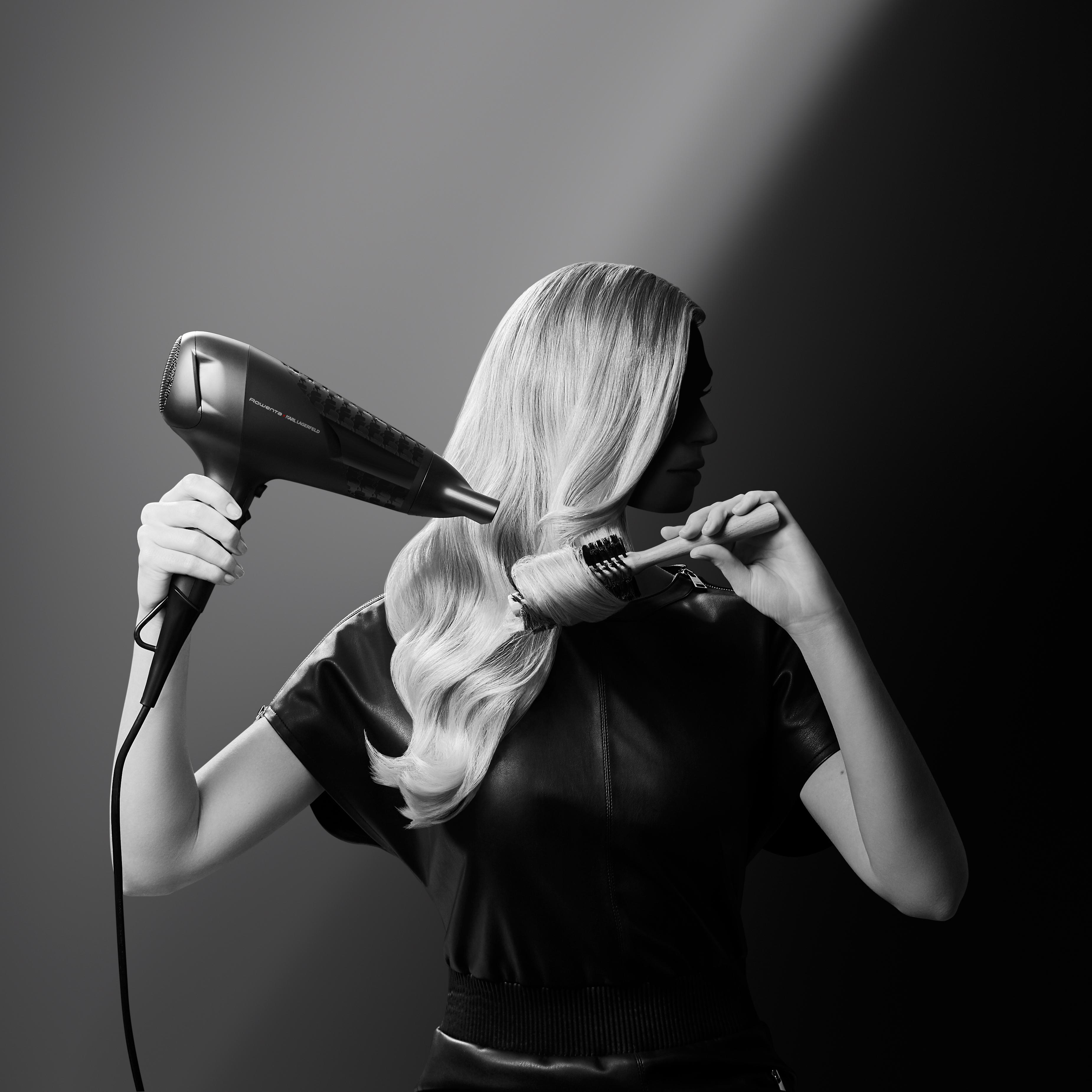 Frau mit blonden Haaren, die sie mit einem Haartrockner und einer Bürste trocknet. Schwarzes Lederoutfit, grauer Hintergrund.