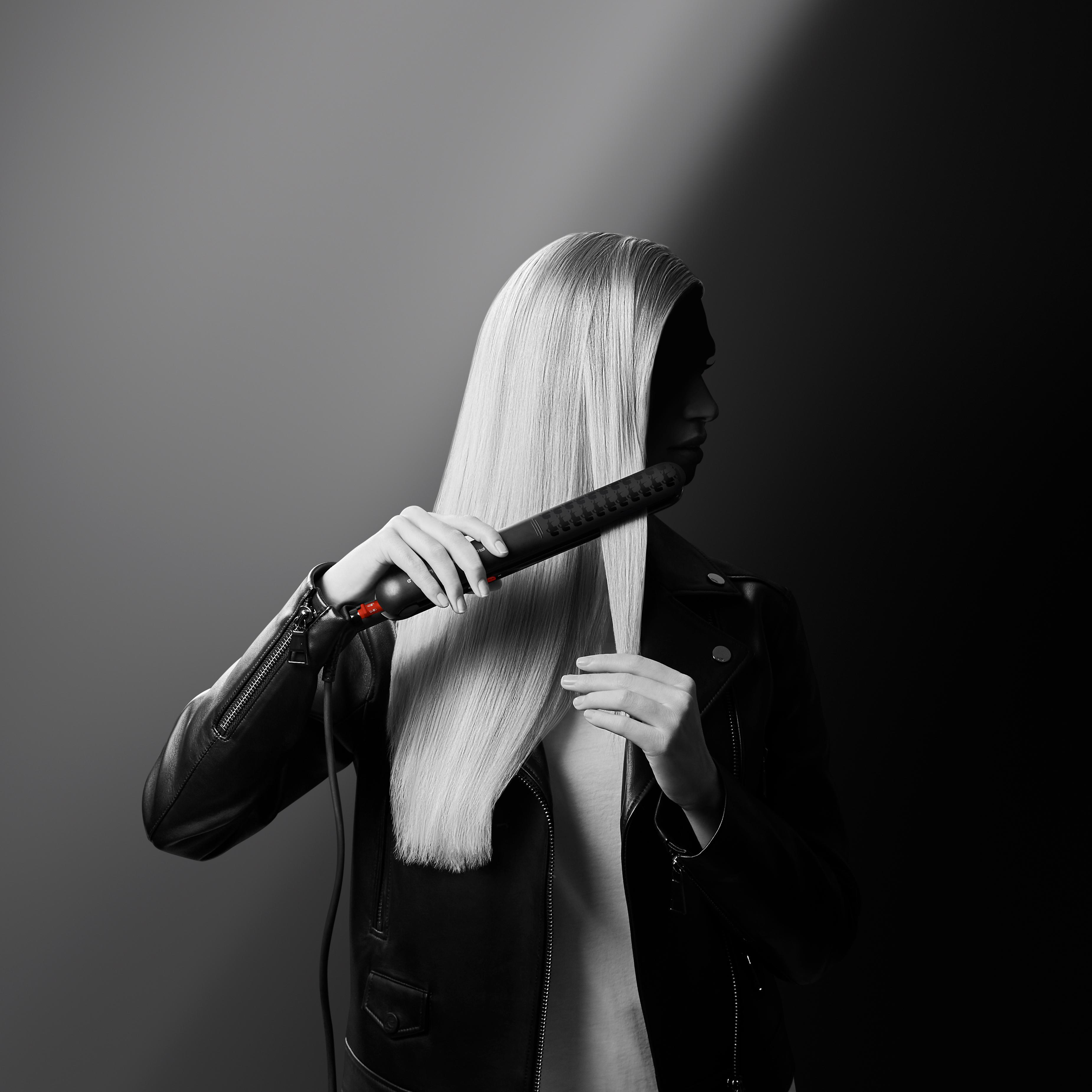 Eine Frau mit langen, blonden Haaren benutzt ein schwarzes Glätteisen. Sie trägt eine schwarze Lederjacke.