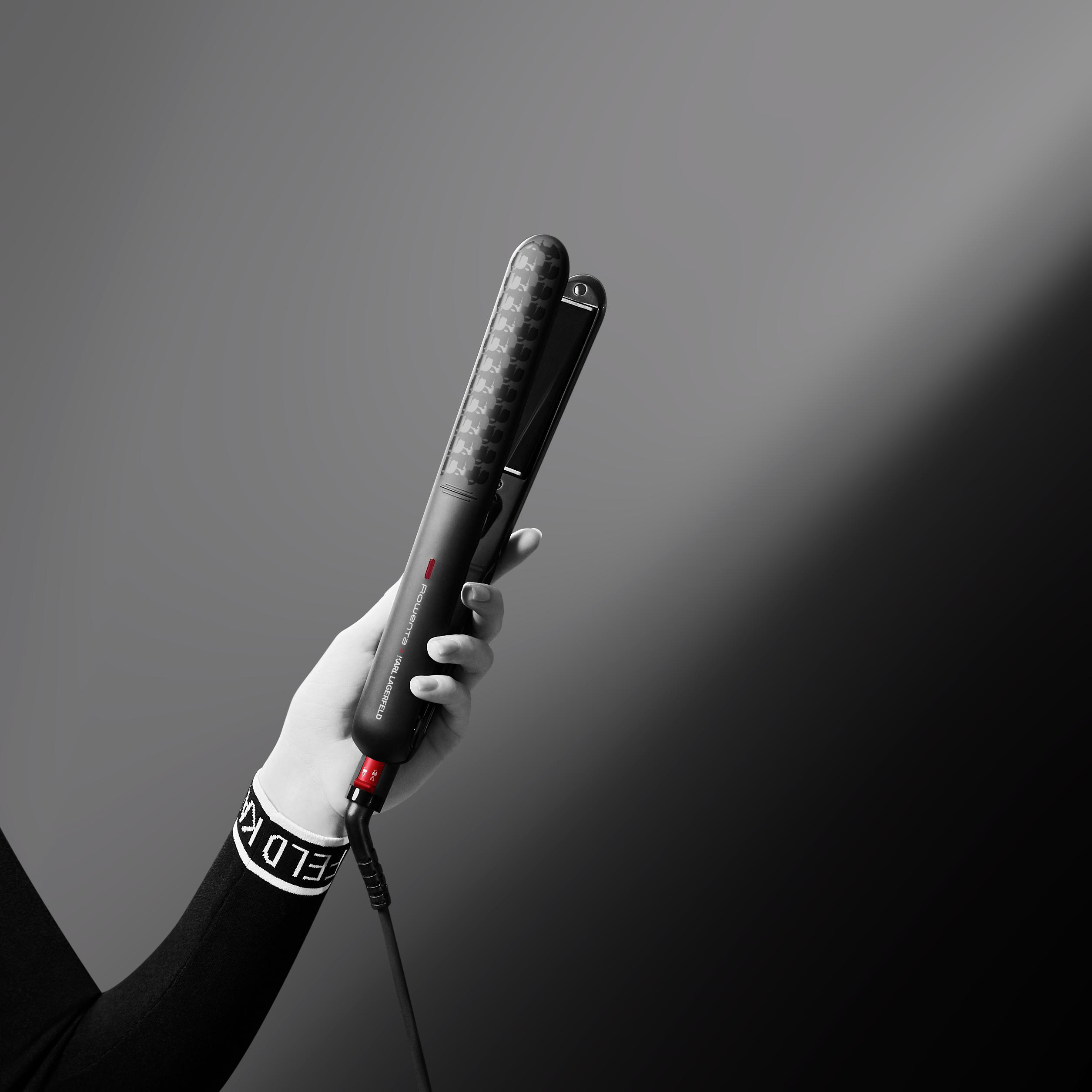 Eine Hand, die einen schwarzen Haarglätter mit roten Akzenten und ein weißes Armband hält.