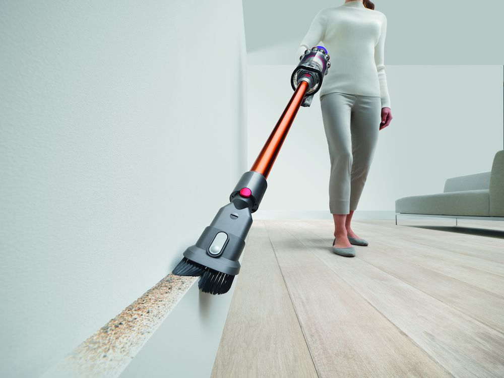 Una persona aspira una esquina de la pared. La aspiradora es naranja y gris. El suelo y la pared son claros.