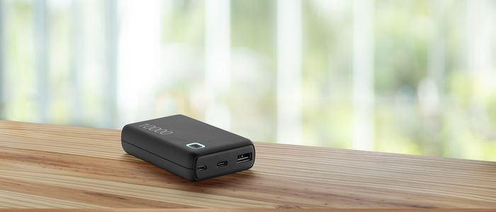 Schwarze Powerbank auf einem Holztisch mit unscharfem Fenster im Hintergrund.