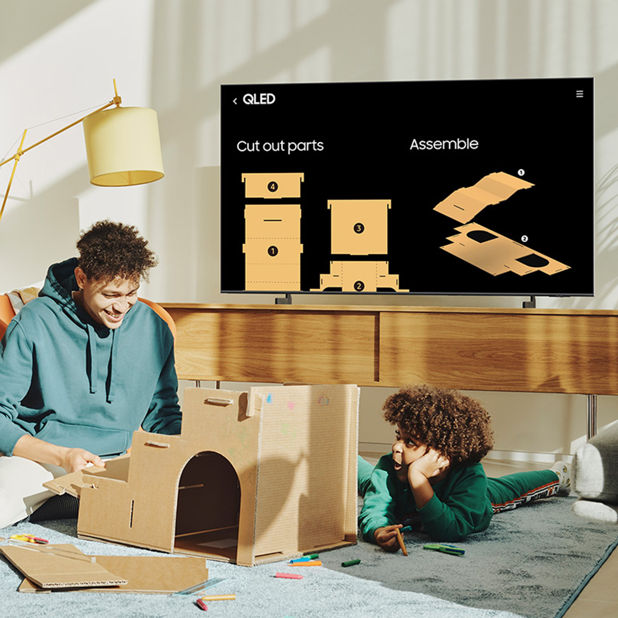 Dos personas construyendo una estructura de cartón frente a un televisor con instrucciones.