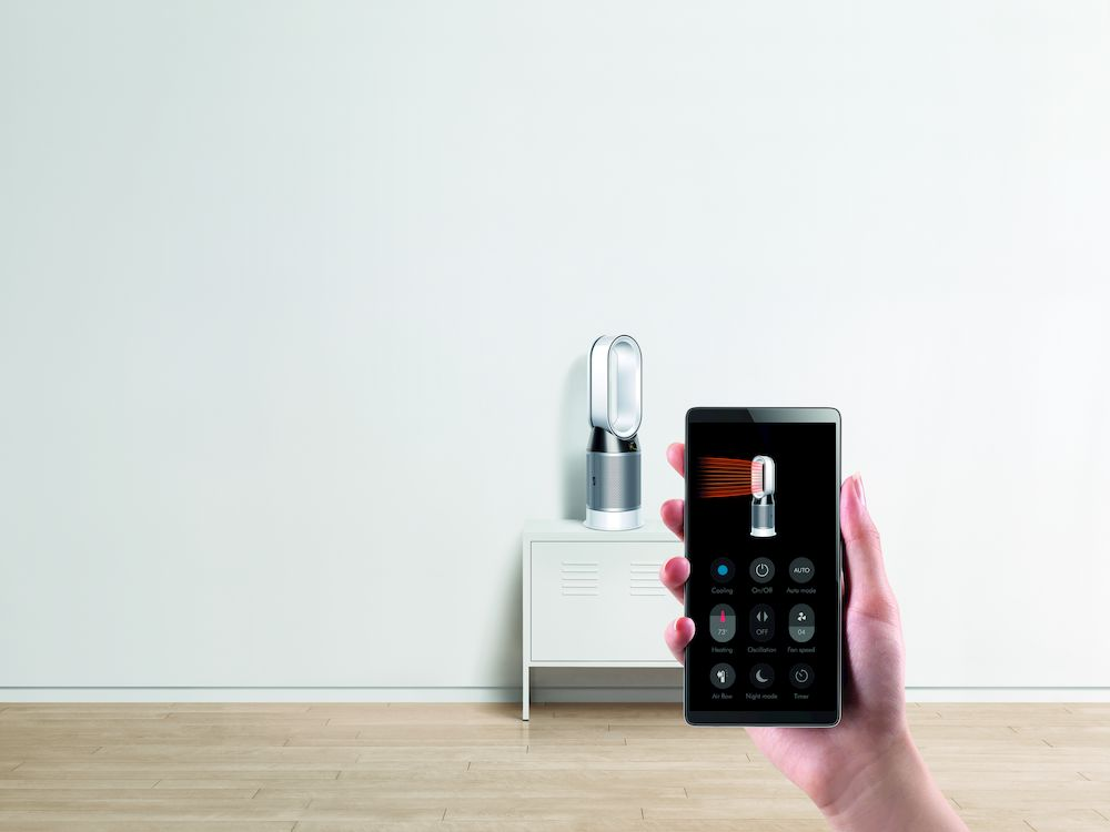 Una mano sosteniendo un smartphone muestra una interfaz de control del ventilador, un ventilador está en un estante blanco.