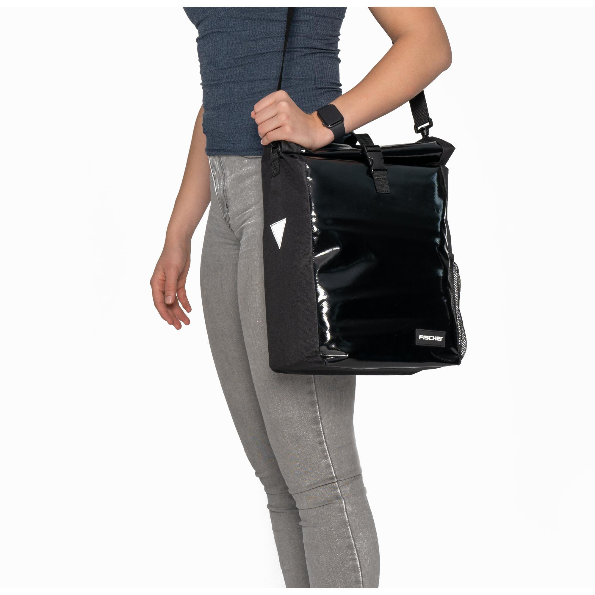 Eine Frau trägt eine schwarze Tasche über der Schulter. Die Tasche ist glänzend und hat eine Klappe. Sie trägt graue Hosen.